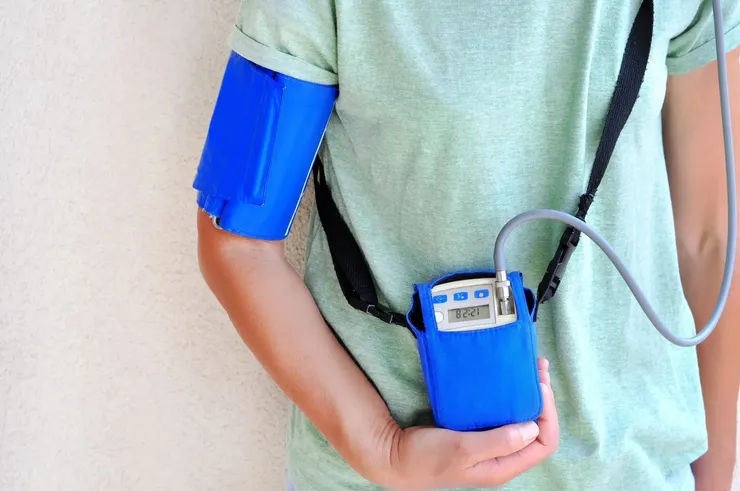Pessoa com braçadeira azul de MAPA no braço, ligada por um tubo ao monitor portátil carregado na bolsa a tiracolo.
